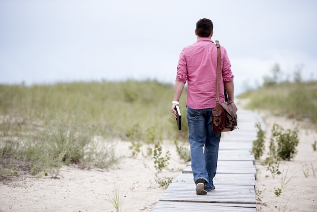 Free photo man walking on a wooden pathway carrying his bag and holding the bible