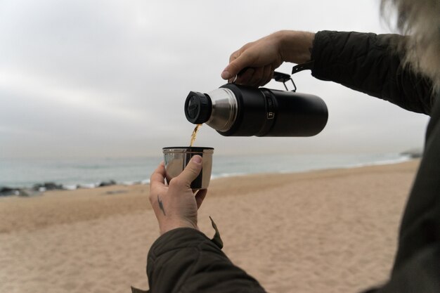 Man in warm jacket with hand tattoos, pours hot coffee or tea into thermos insulated cup on cold rainy day.