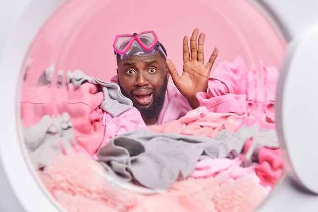  man waves palm does laundry at home wears snorkeling mask photographed from inside of washing machine Afro American householder going to wash dirty clothes occupied with household