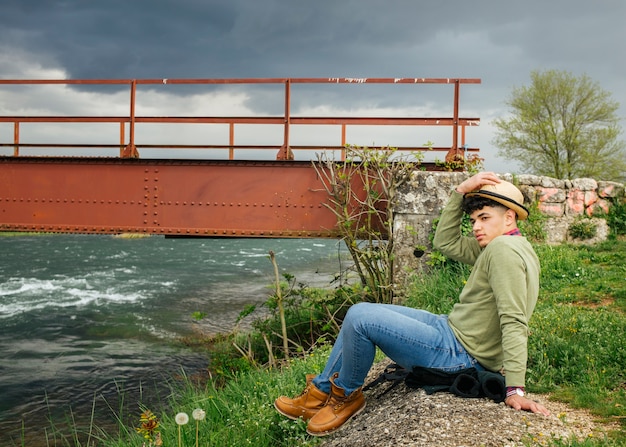 Foto gratuita cappello da portare dell'uomo che si siede vicino al fiume del fiore