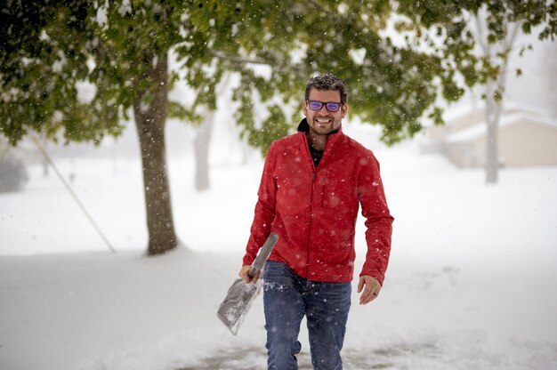 Man wearing a red jacket and walking in a snowy field while holding the snow shovel