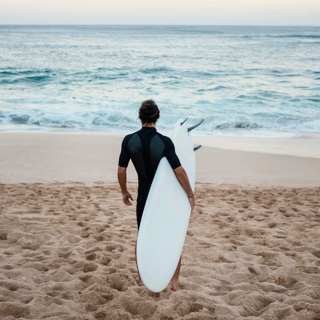 Foto gratuita uomo che indossa abiti da surfista che cammina sulla sabbia da dietro