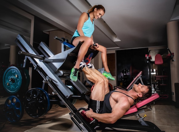 Man weightlifter doing leg presses with his trainer. Sports couple is working out at gym.