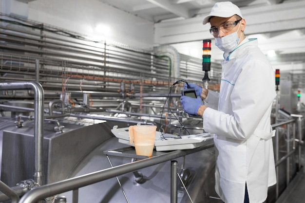 Man in a white coat gloves and a mask stands near the production line and holds a tester with wires at the dairy plant The inspector carries out control at the cheese factory