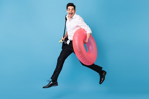 Free photo man in white shirt and black trousers runs against blue space, happily smiles and holds inflatable circle.