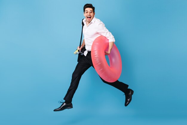 Man in white shirt and black trousers runs against blue space, happily smiles and holds inflatable circle.