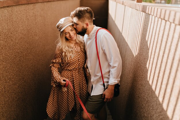 Man in white shirt and khaki shorts gently kisses his positive smiling blonde wife in brown dress.