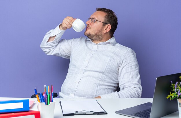 青のラップトップとオフィスのフォルダーとテーブルに座って自信を持ってお茶を飲む眼鏡をかけている白いシャツの男