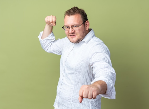 Uomo in camicia bianca con gli occhiali che guarda davanti con la faccia seria che stringe i pugni in piedi sul muro verde