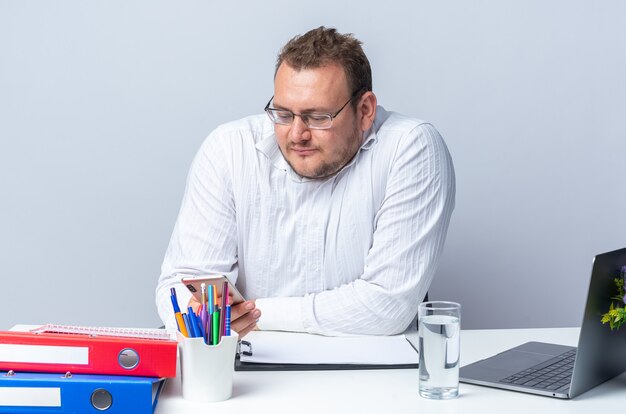 피곤하고 지루해 보이는 안경을 쓴 흰 셔츠를 입은 남자가 사무실에서 일하는 흰 벽 위에 노트북 사무실 폴더와 클립보드를 들고 테이블에 앉아 있다