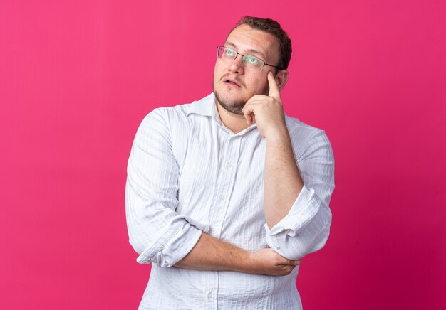 Человек в белой рубашке в очках смотрит озадаченно, стоя на розовом