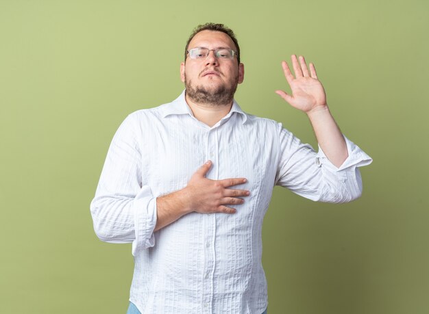 Man in white shirt wearing glasses  taking an oath raising hand with other hand on his chest  with serious face standing over green wall