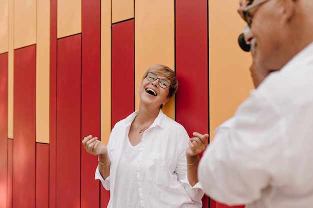 Man in white stylish shirt photographing blonde cheerful woman with eyeglasses in light blouse on red and orange.