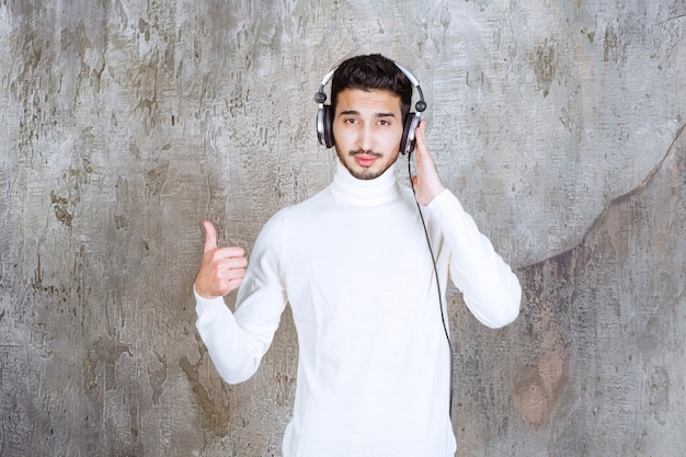 Человек в белом свитере в наушниках, слушающий музыку и показывающий положительный знак рукой