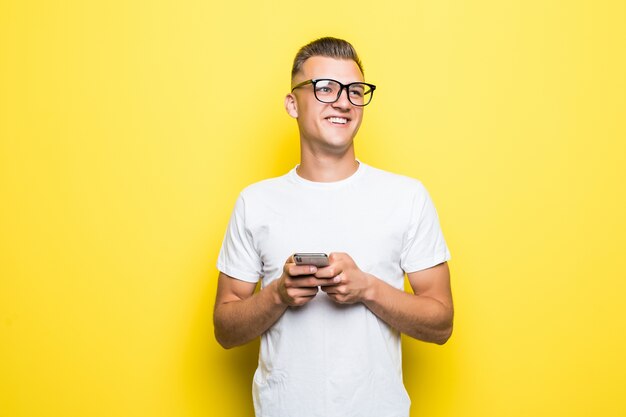 Man in white t-shirt and glasses makes something on his phone and takes selfie pictures isolated on yellow