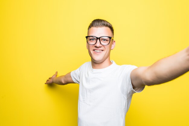 Man in white t-shirt and glasses makes something on his phone and takes selfie pictures isolated on yellow