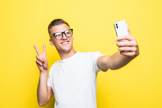 Man in white t-shirt and glasses makes something on his phone and takes selfie pictures victory sign