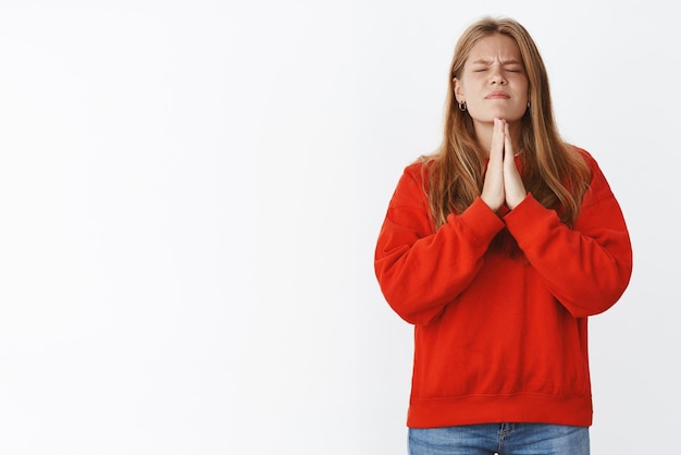 Man wishing for dream come true, holding hands in pray focusing on prayers, closing eyes and faithfully hoping for god help and fortune come, posing in cute warm onsized sweater on grey wall.