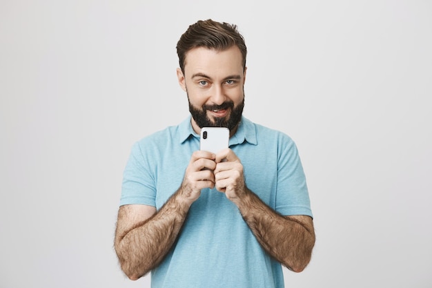 Бесплатное фото Человек с бородой держит белый смартфон с забавным выражением лица