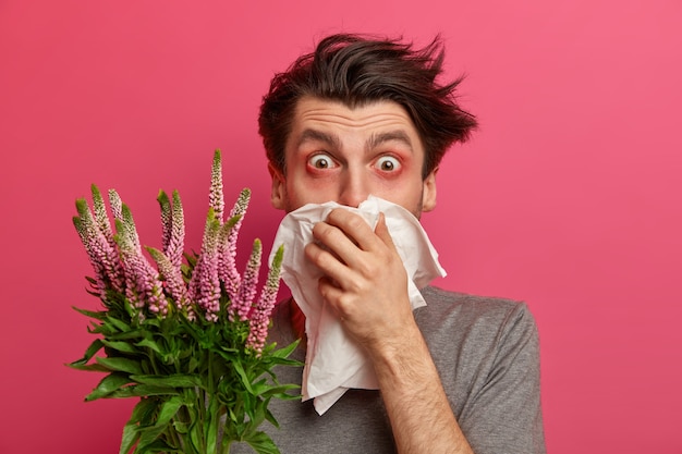 Foto gratuita l'uomo con allergia starnutisce e copre il naso con un tovagliolo, ascolta i consigli dell'allergologo su come curare la febbre da fieno, ha gli occhi rossi e lacrimosi, ha bisogno di trattare la rinite allergica, isolato sul muro rosa.