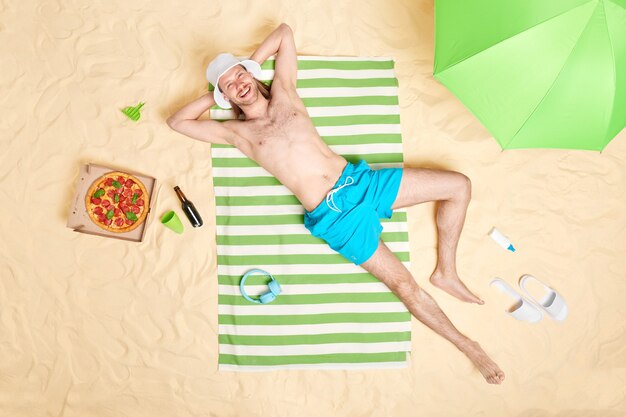  man with bare torso smiles gladfully wears sunhat and blue shorts poses topless on striped towel surrounded by beach accessories has lazy day good rest at seaside. Summer time concept