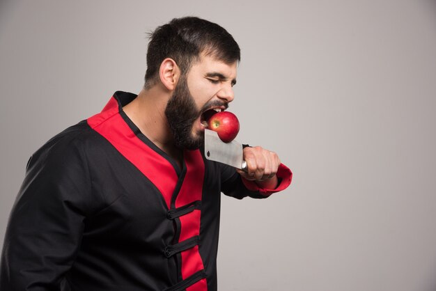 Man with beard biting a red apple on knife .