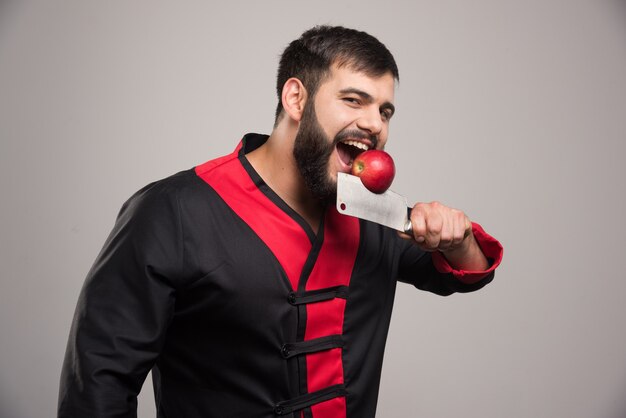 Man with beard biting a red apple on knife .