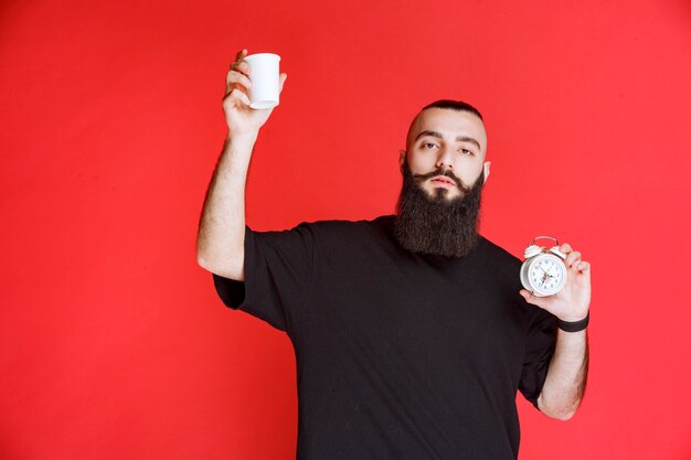 Мужчина с бородой держит будильник и чашку кофе.