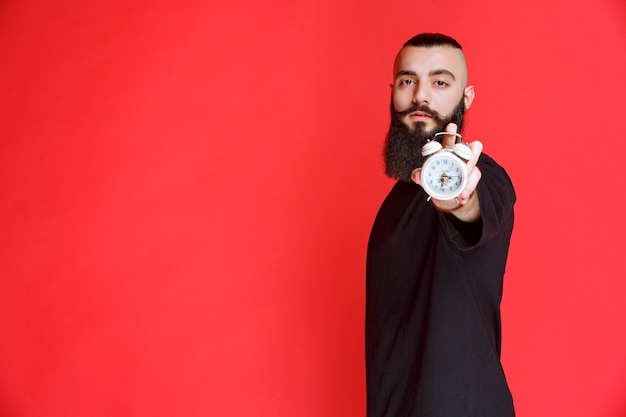 Foto gratuita uomo con la barba che tiene e promuove una sveglia come prodotto.