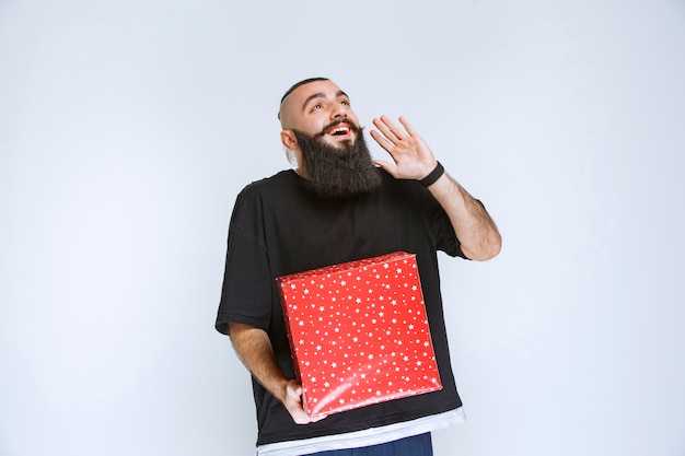 Man with beard holding a red gift box and calling someone or whispering.  