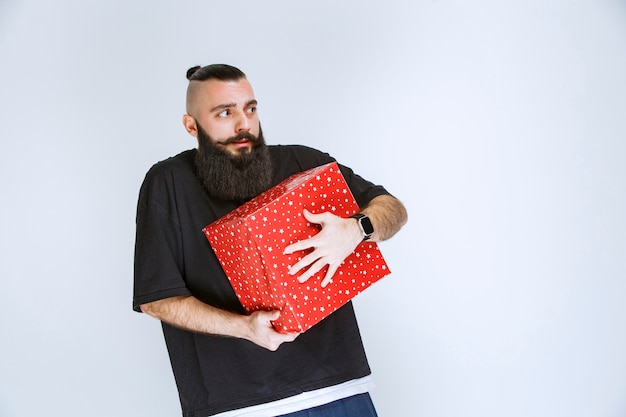 Man with beard holding a red gift box and looks confused and terrified.  