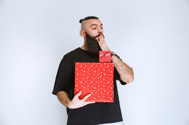 Man with beard holding a red gift box and looks confused and terrified.  