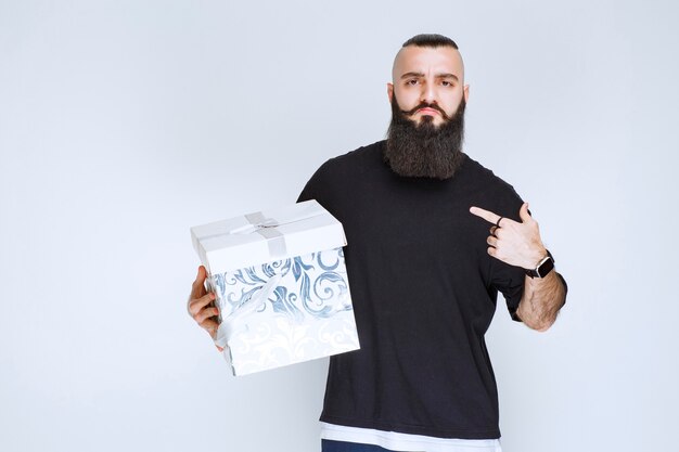 Man with beard holding a white blue gift box and demonstrating it. 