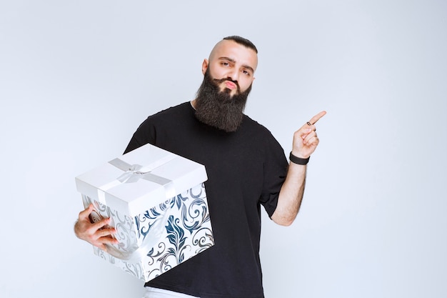 Man with beard holding a white blue gift box and demonstrating it. 