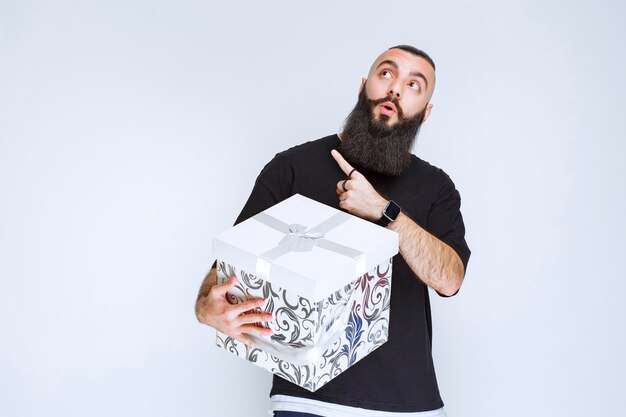 Foto gratuita uomo con la barba che tiene in mano una confezione regalo blu bianca e lo dimostra.