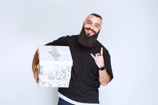 Мужчина с бородой держит бело-синюю подарочную коробку и чувствует себя успешным.