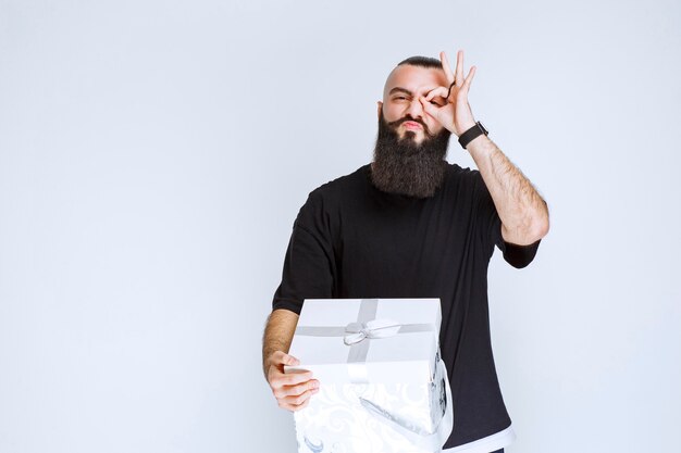 Man with beard holding a white blue gift box and looking across his fingers. 