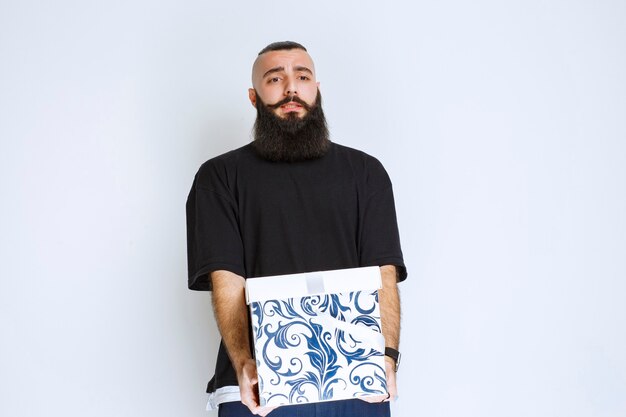 Man with beard holding a white blue gift box and looks disappointed. 