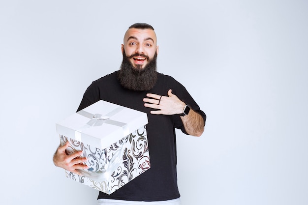 Man with beard holding a white blue gift box and looks surprized. 