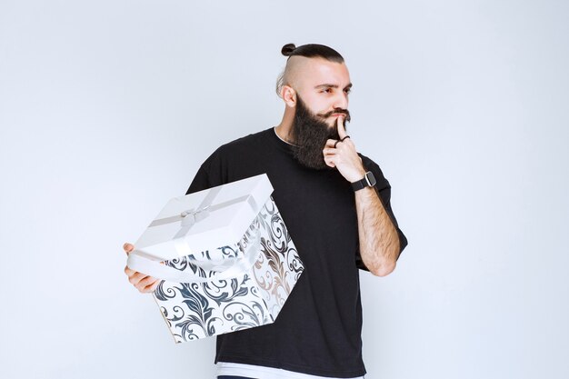 Man with beard holding a white blue gift box pointing his mouth and asking for silence. 