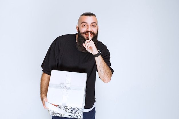 Man with beard holding a white blue gift box pointing his mouth and asking for silence. 