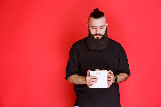 Man with beard holding a white gift box and looks doubtful about what is inside.