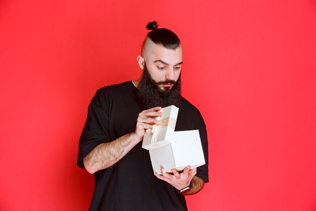 Man with beard holding a white gift box and looks doubtful about what is inside.