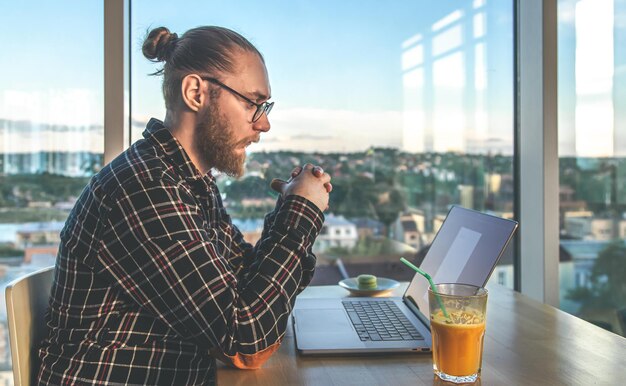 あごひげを生やした男性が、窓際のオフィスに座っているコンピューターで働いています。