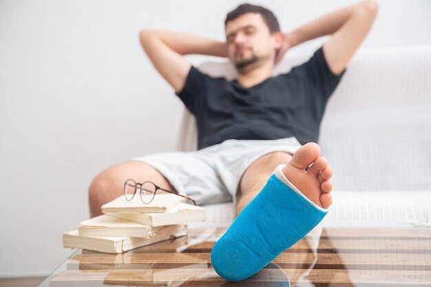 Мужчина со сломанной ногой в синей шине для лечения травм от растяжения связок голеностопного сустава читает книги в домашней реабилитации.