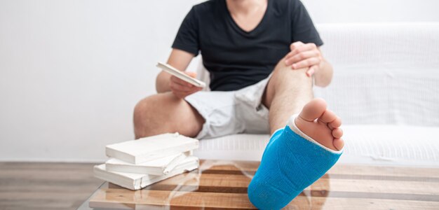 Мужчина со сломанной ногой в синей шине для лечения травм от растяжения связок голеностопного сустава читает книги в домашней реабилитации.