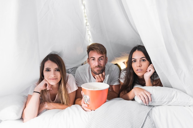 Man with bucket of popcorns lying on bed with two female friends
