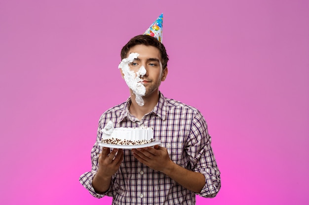 Foto gratuita uomo con la torta sul viso sopra il muro viola. festa di compleanno.