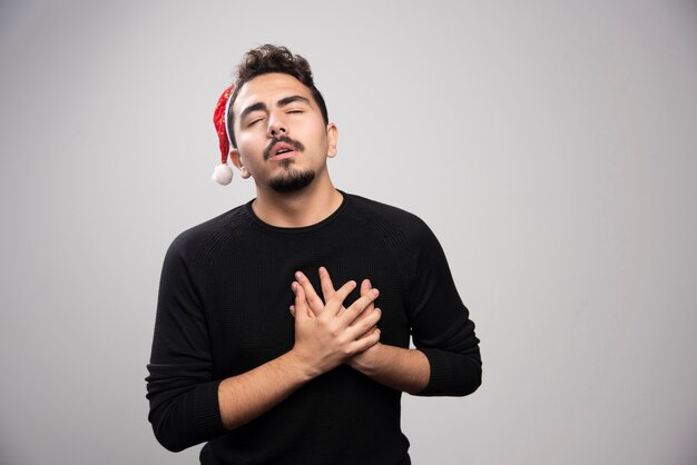 Man with closed eyes in Santa Claus red hat put his hands on his heart. 