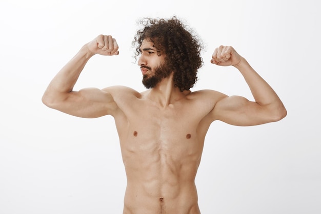 Free photo man with curly hair looking left and raising arms working out and being satisfied with own body standing naked